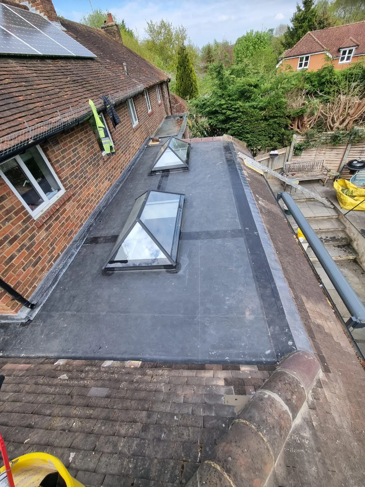 Grey fibreglass flat roof with two roof lanterns beside a tiled brick house